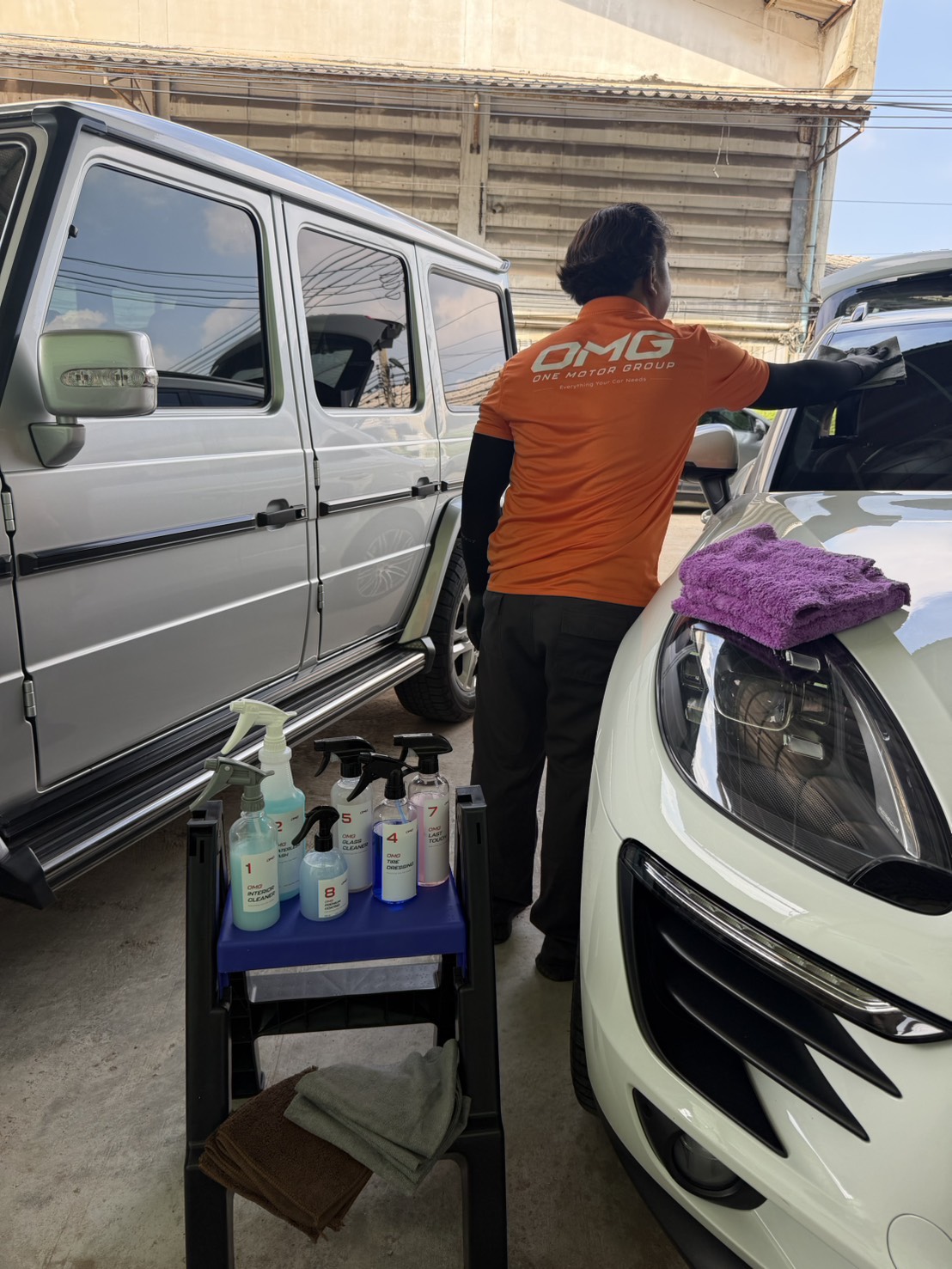 OMG technician in an orange uniform wiping a white SUV windscreen beside a cart of numbered OMG waterless products