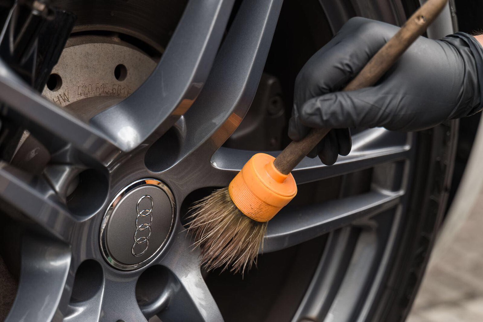 Gloved hand detailing an alloy wheel with an orange-handled brush